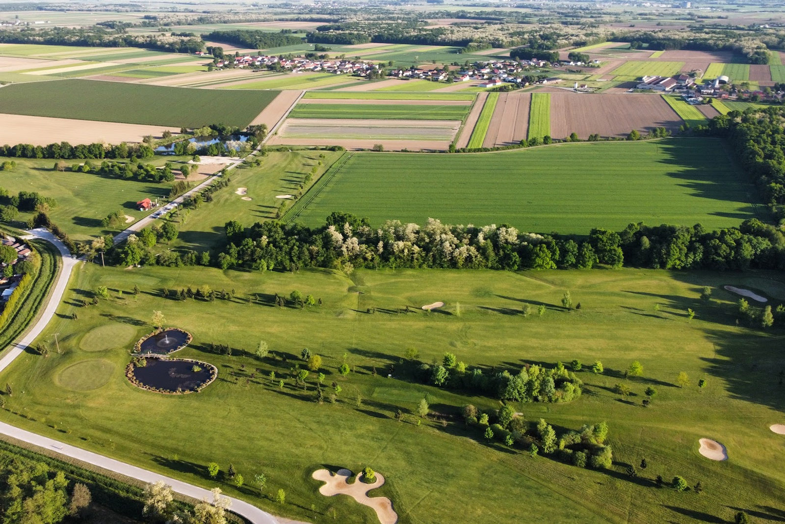 Drohnenaufnahme aus der Luft von einem grünen Golfplatz mit Teichen und Bunkern, umgeben von einer Flickenteppich-Landschaft und einem Dorf am Horizont