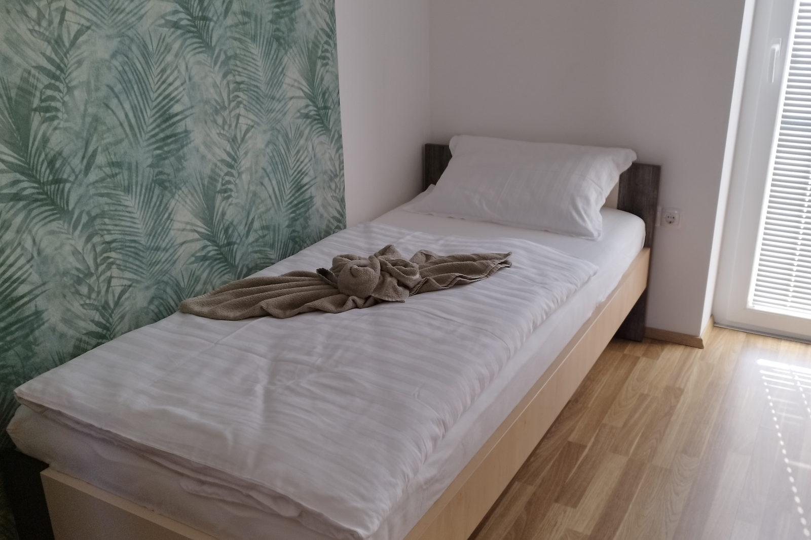 Einzelbett mit weißer Bettwäsche und einem gefalteten beigen Handtuch, an einer Wand mit grüner Palmblatt-Tapete, neben einer Balkontür