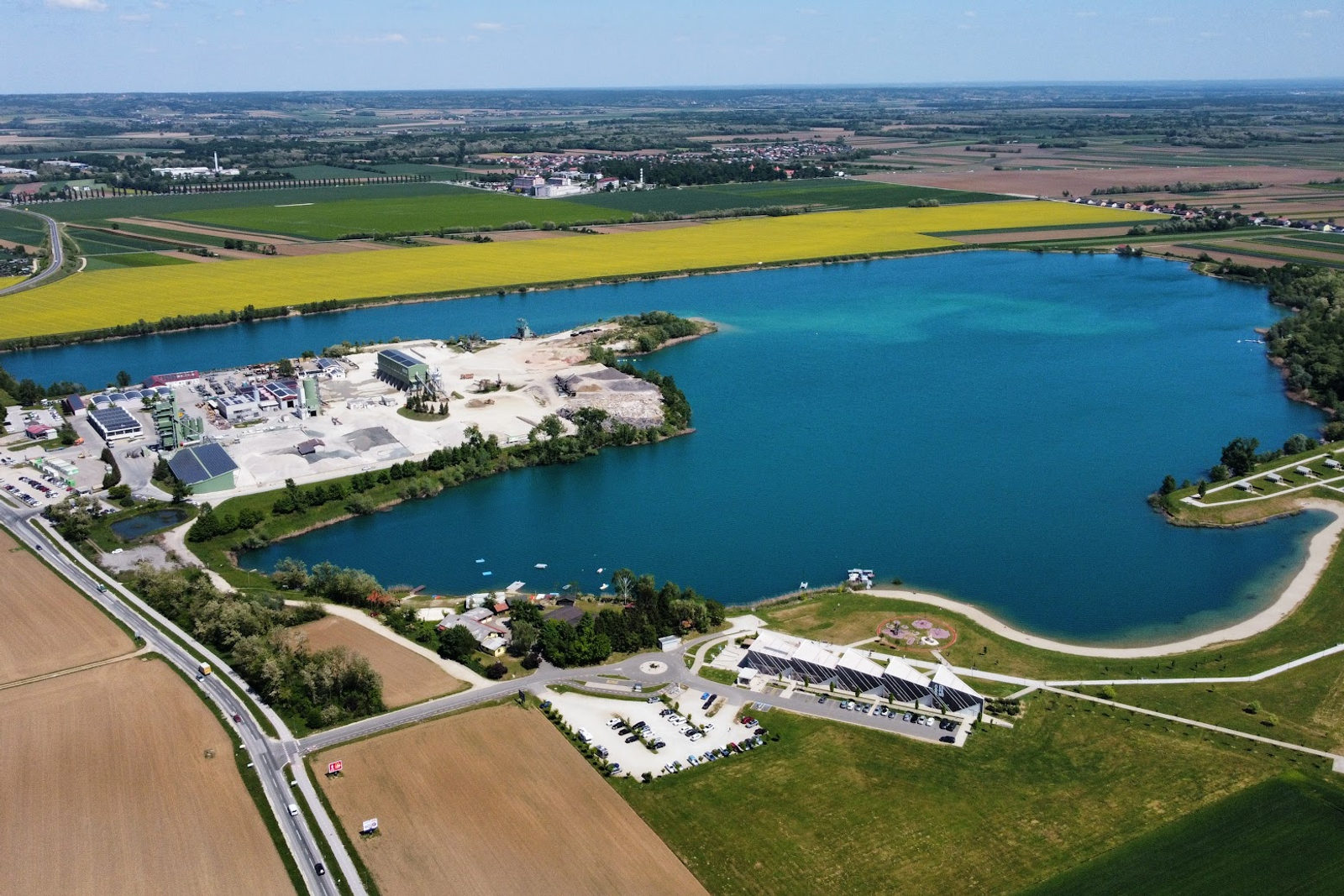 Drohnenaufnahme aus der Luft von einem großen türkisfarbenen See, umringt von Feldern, mit Sandstrand, einem Seepavillon und einem gelben Rapsfeld dahinter