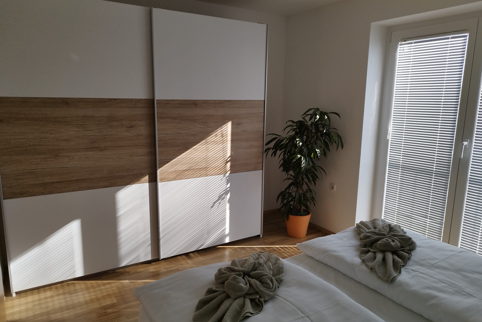 Schlafzimmer mit großem weiß-eichenem Schiebeschrank, einer Topfpflanze an der Balkontür und einem Doppelbett mit gefalteten Handtüchern im Sonnenlicht