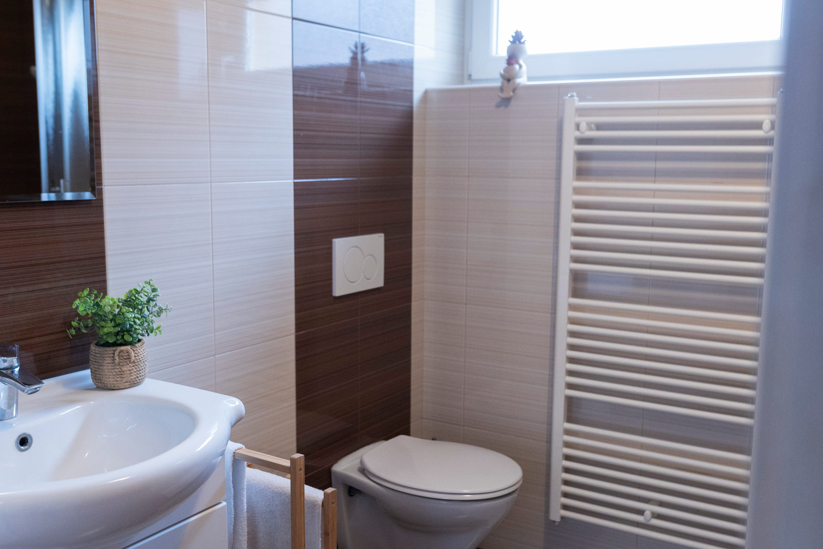 Badezimmer mit weißem Waschbecken, einer kleinen grünen Pflanze, einer wandhängenden Toilette und einem weißen Handtuchheizkörper vor gestreiften braunen Fliesen