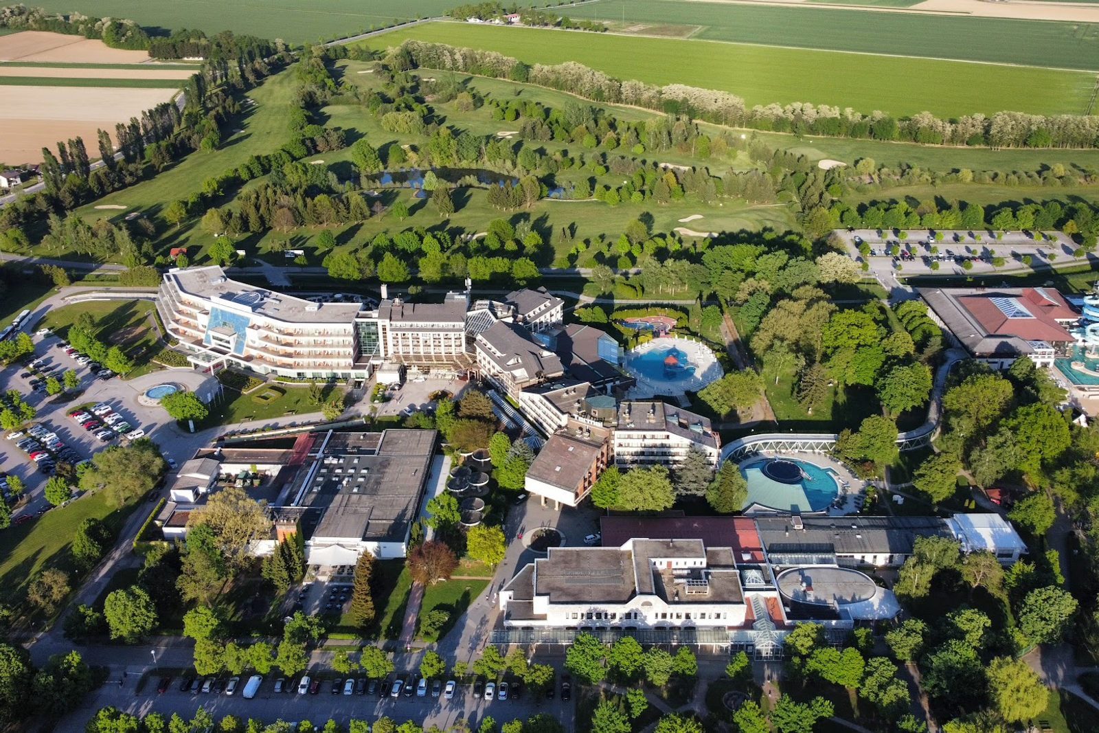 Luftaufnahme eines Thermenresorts mit einem geschwungenen weißen Hotel, Thermalbecken im Freien, alten Bäumen, Parkplätzen und einem Golfplatz dahinter
