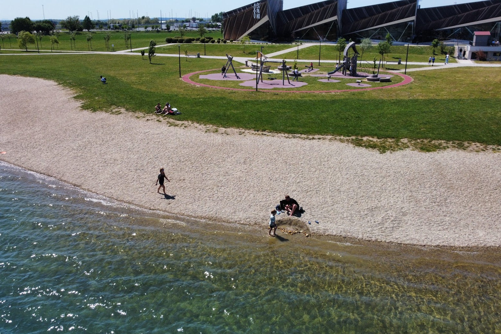 Kiesstrand an einem klaren See mit einigen Badenden, einer Wiese, einem Kinderspielplatz und dem kantigen hölzernen Expano-Pavillon dahinter
