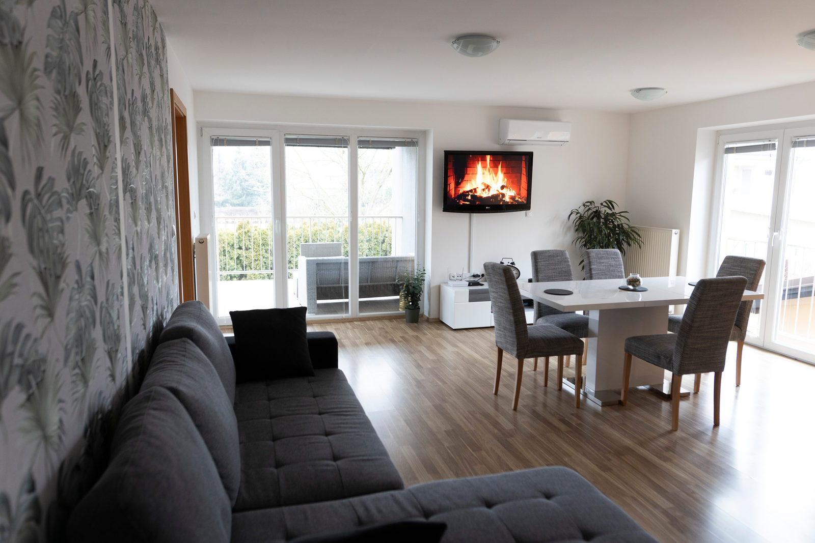Helles Wohnzimmer mit grauer Ecksofa-Garnitur, blattgemusterter Tapete, weißem Esstisch mit sechs Stühlen, einem Wandfernseher und Türen zur Terrasse