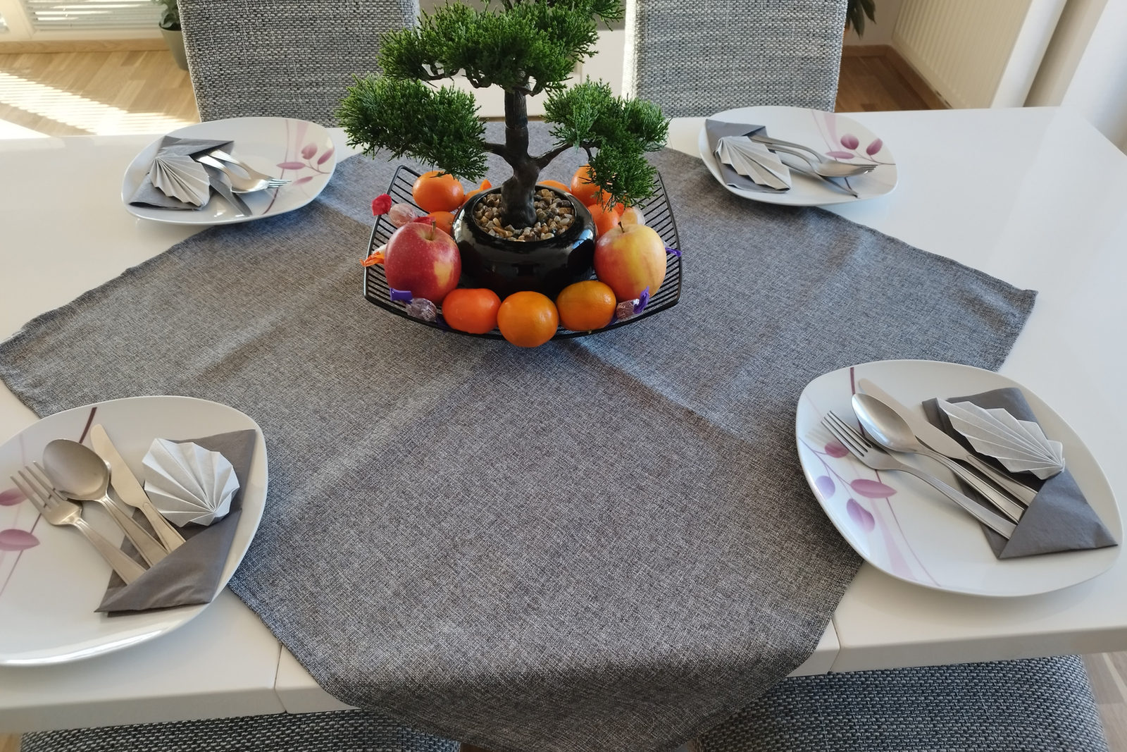 Weißer Esstisch für vier gedeckt, mit gefalteten Servietten, Besteck und einer schwarzen Schale mit Äpfeln und Mandarinen um einen kleinen Bonsai