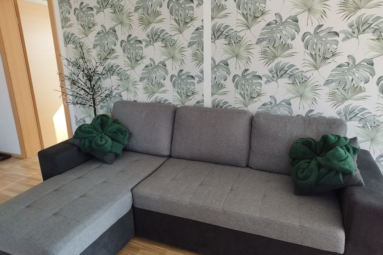 Graue Ecksofa-Garnitur mit dunkelgrün gefalteten Decken auf den Armlehnen, vor weißer Tapete mit großen Monstera- und Palmblättern